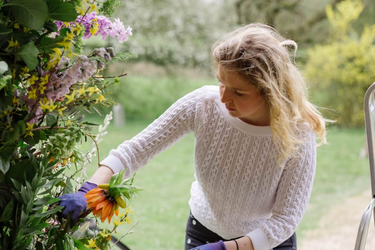 Kirsty arranges wedding flowers outdoors, placing an orange bloom into a lush display of mixed greenery and seasonal flowers. Soft natural light and a garden setting highlight the organic, just-picked style of the floral design.