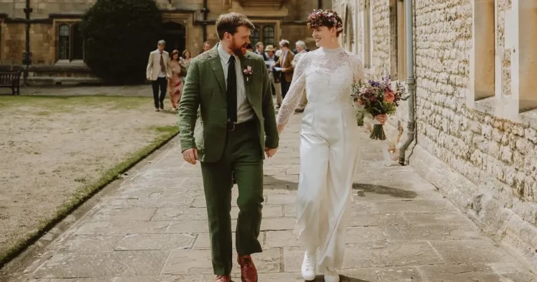 Couple walks hand in hand outside a historic stone building, with one holding a bouquet of wedding flowers in rich, natural tones. Guests follow behind as the scene captures a relaxed moment after the ceremony in a classic courtyard setting.