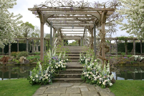 Garden ceremony setup with wedding flowers arranged along stone steps leading to a wooden pergola covered in climbing greenery and blossoms. Soft white and pastel floral arrangements frame the aisle beside a reflective pond, creating a romantic outdoor wedding setting.